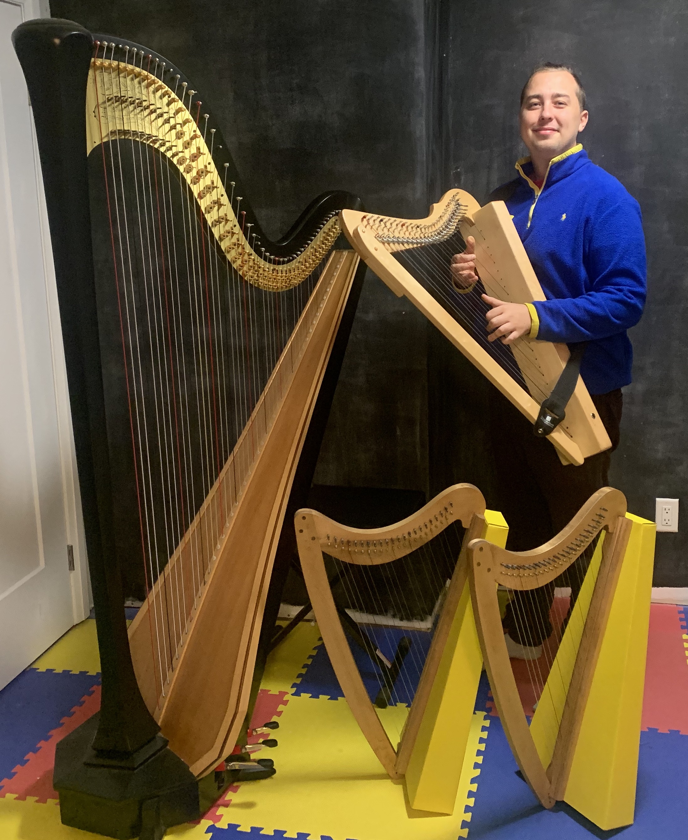 A photo of Andrew infront of a blackboard wearing a small harp and blue sweater, beside a concert grand harp, with two more harps standing on the floor. The floor is colourful soft tiles.